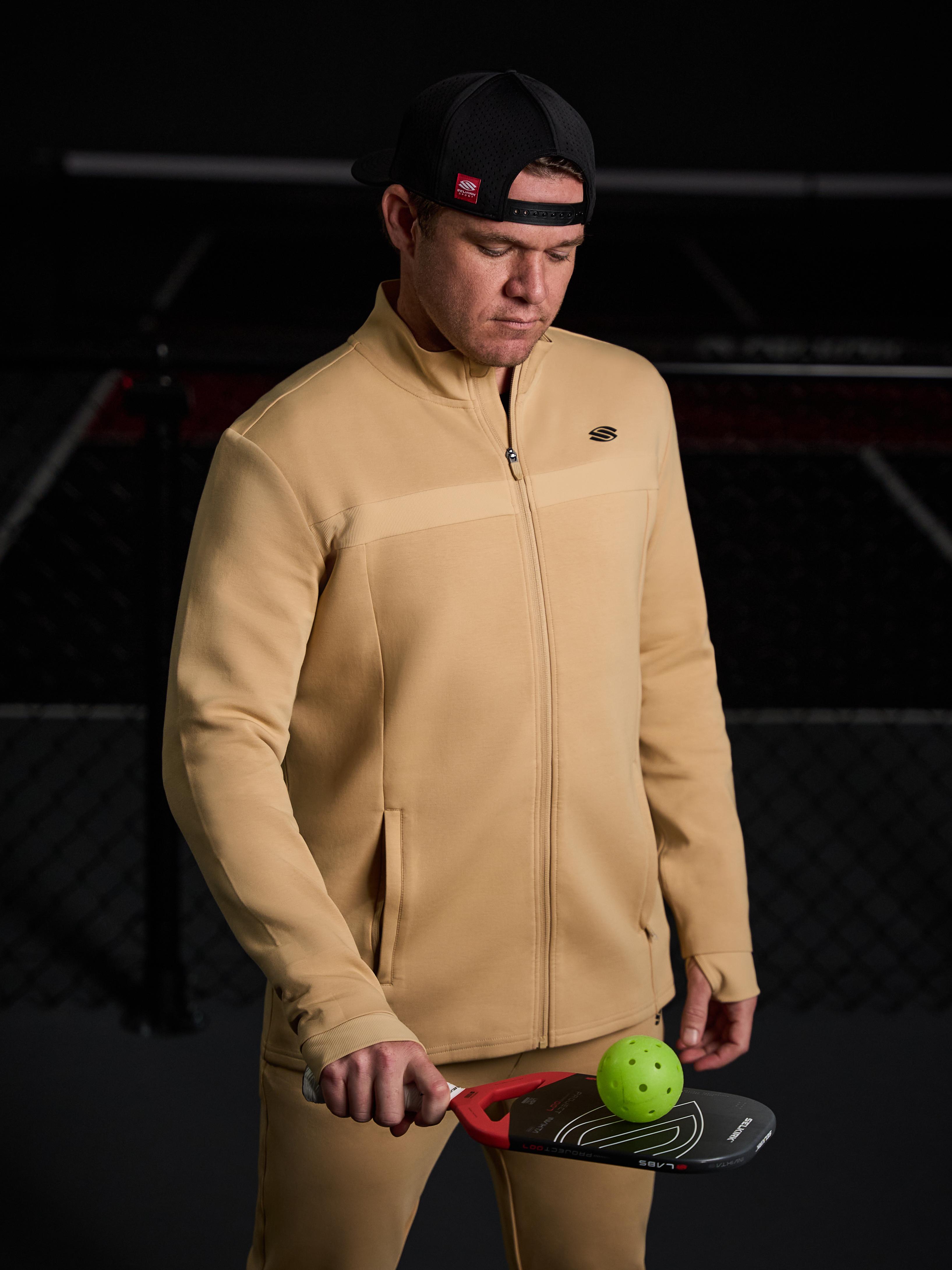 Man wearing a beige full zip jacket, holding a pickleball paddle and ball.