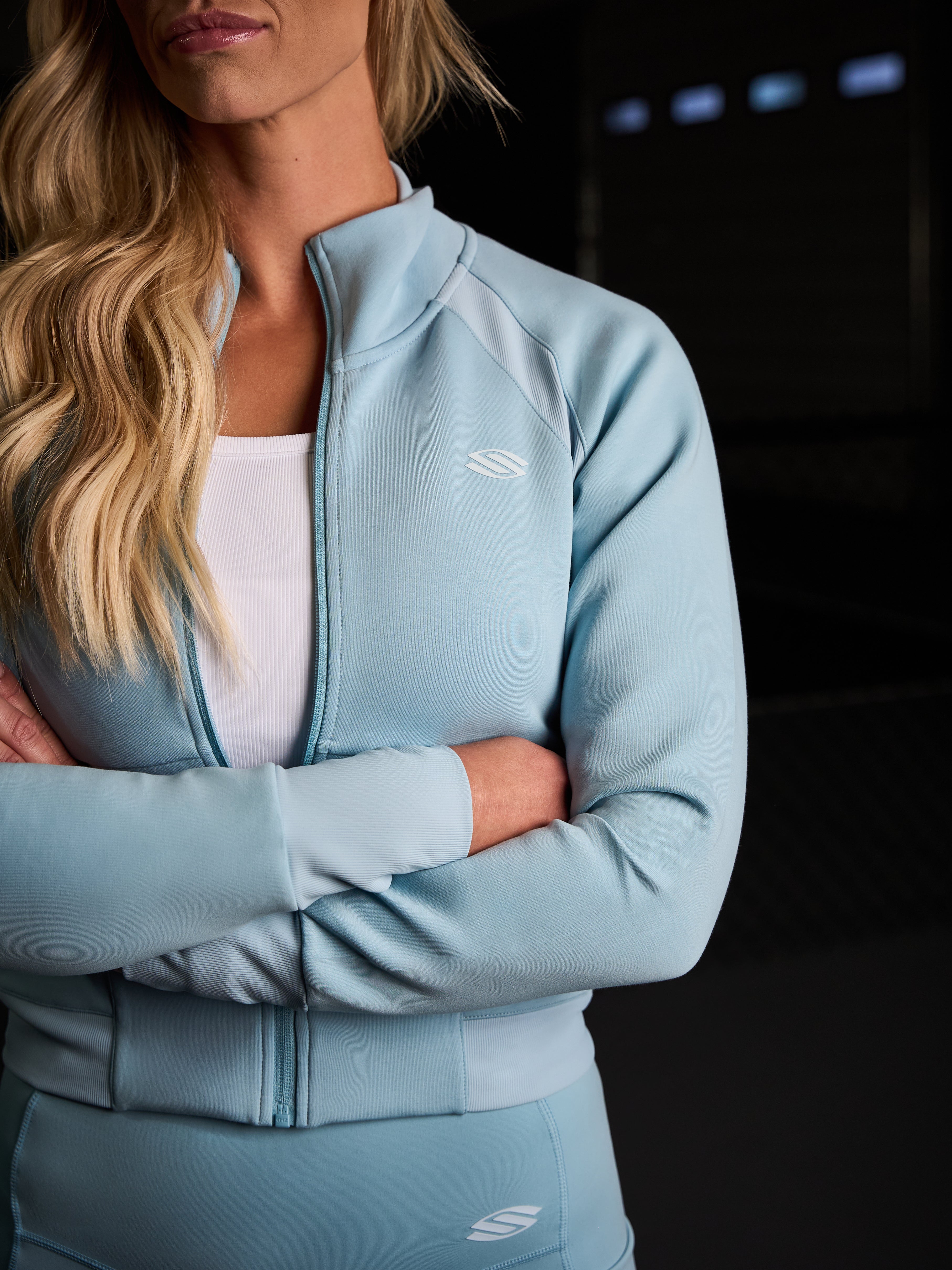 Model wearing a light blue cropped zip-up jacket with long sleeves, arms crossed, showcasing the jacket's fit and style.