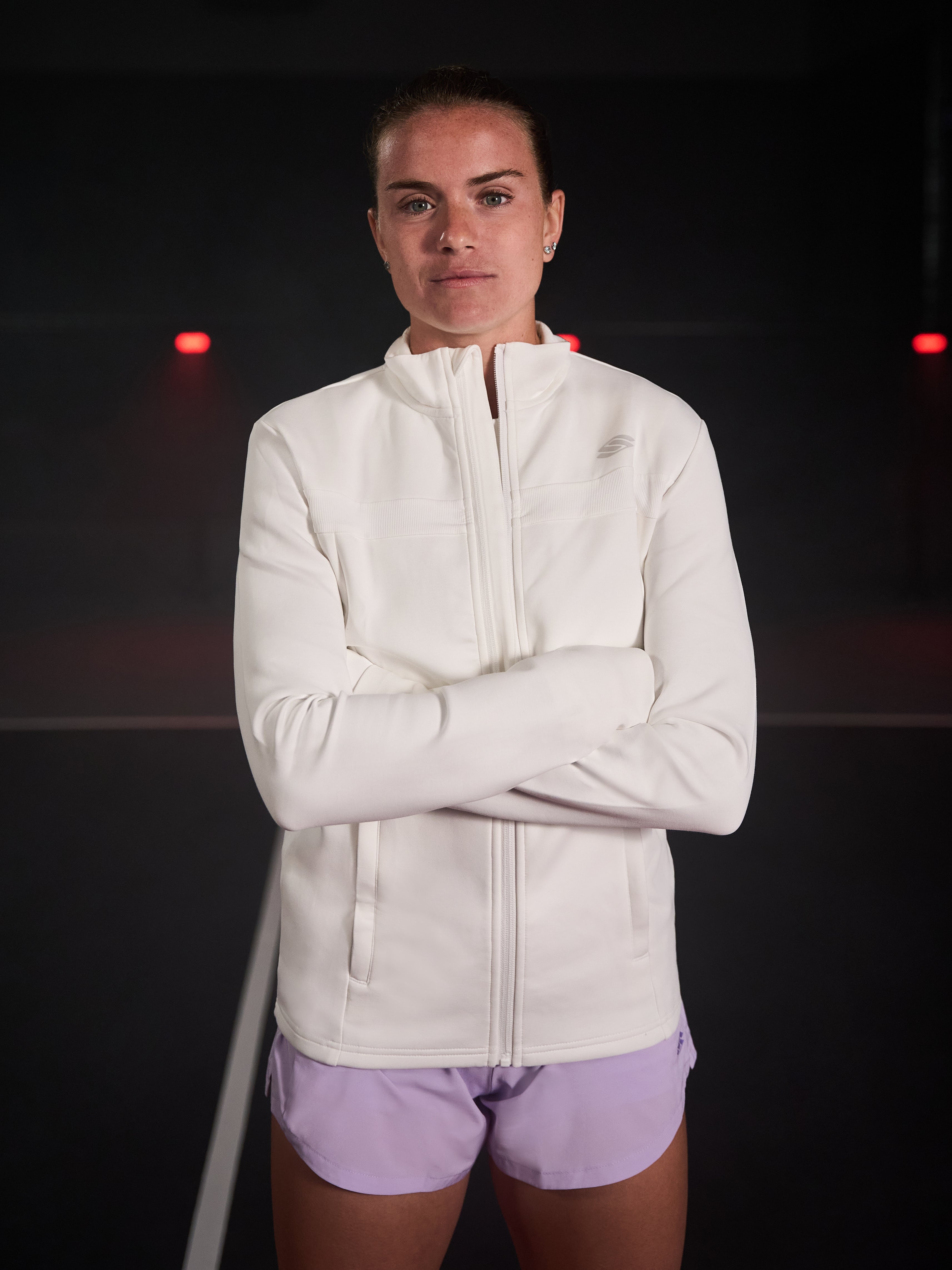 Model wearing a white cropped zip-up jacket, arms crossed, on a court