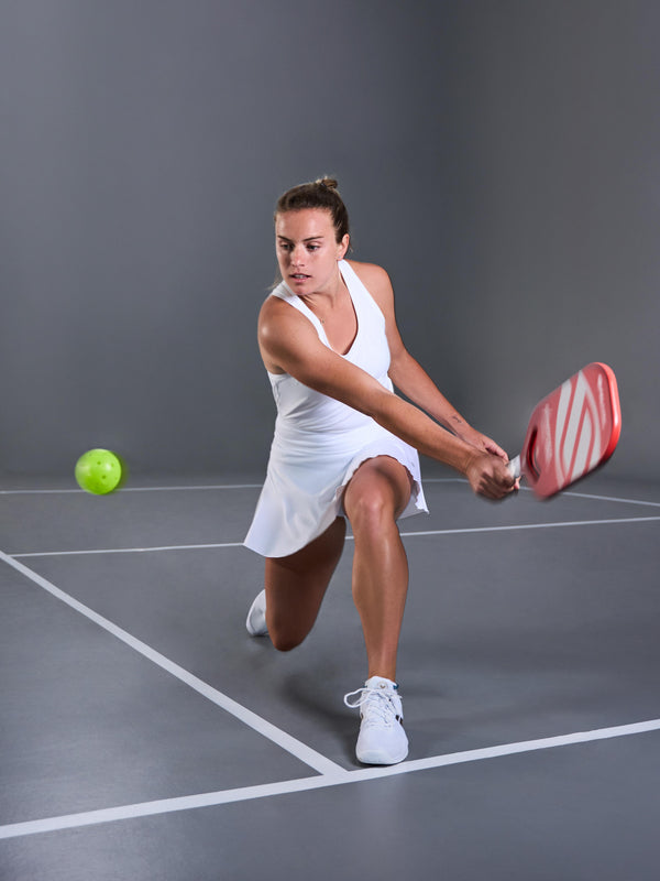 Female player in white dress hitting a pickleball with AMPED Pro Air paddle in red.