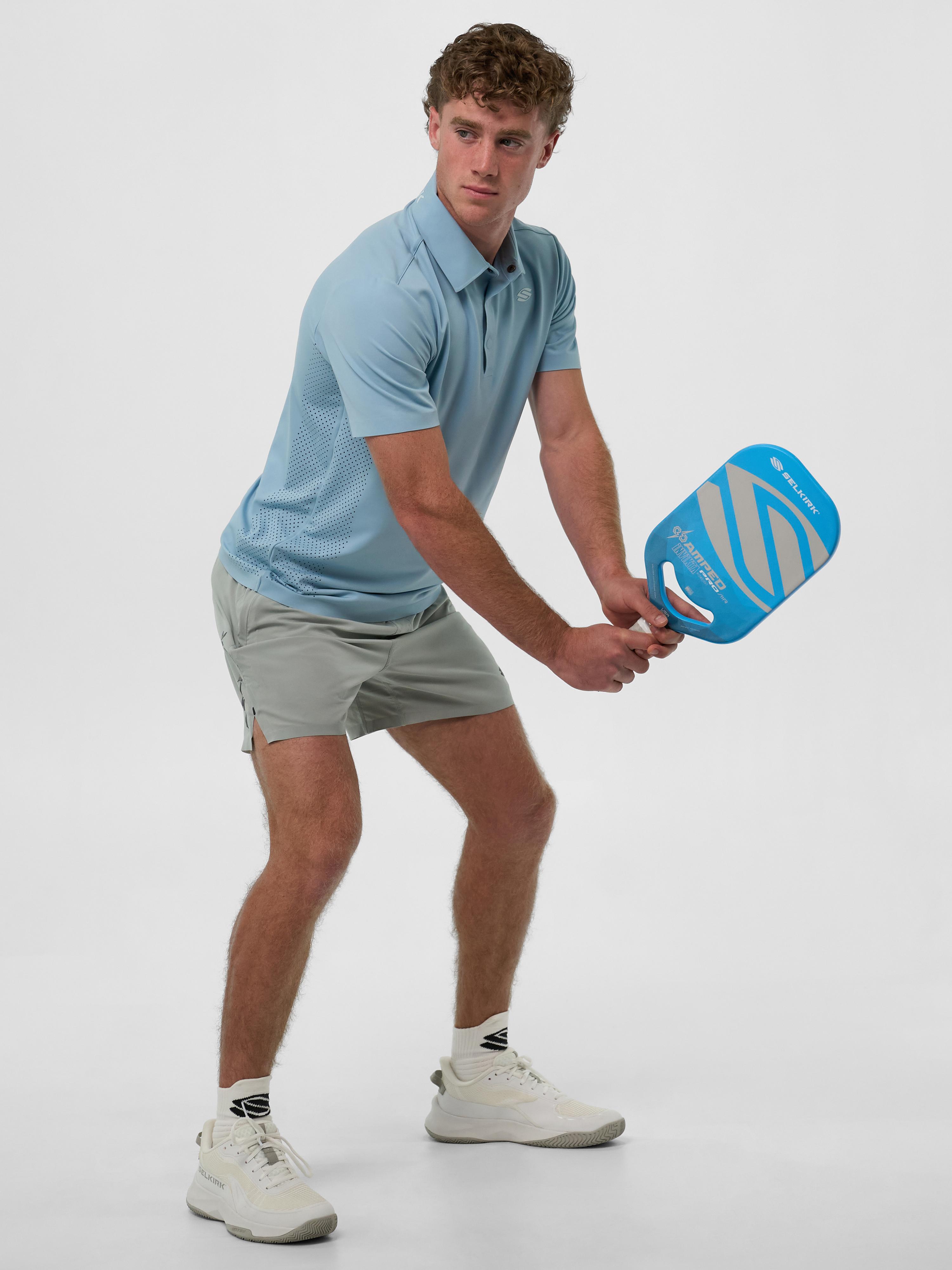 Model wearing a light blue Men's Pro Line Performance Polo, preparing to hit a pickleball with a blue paddle.