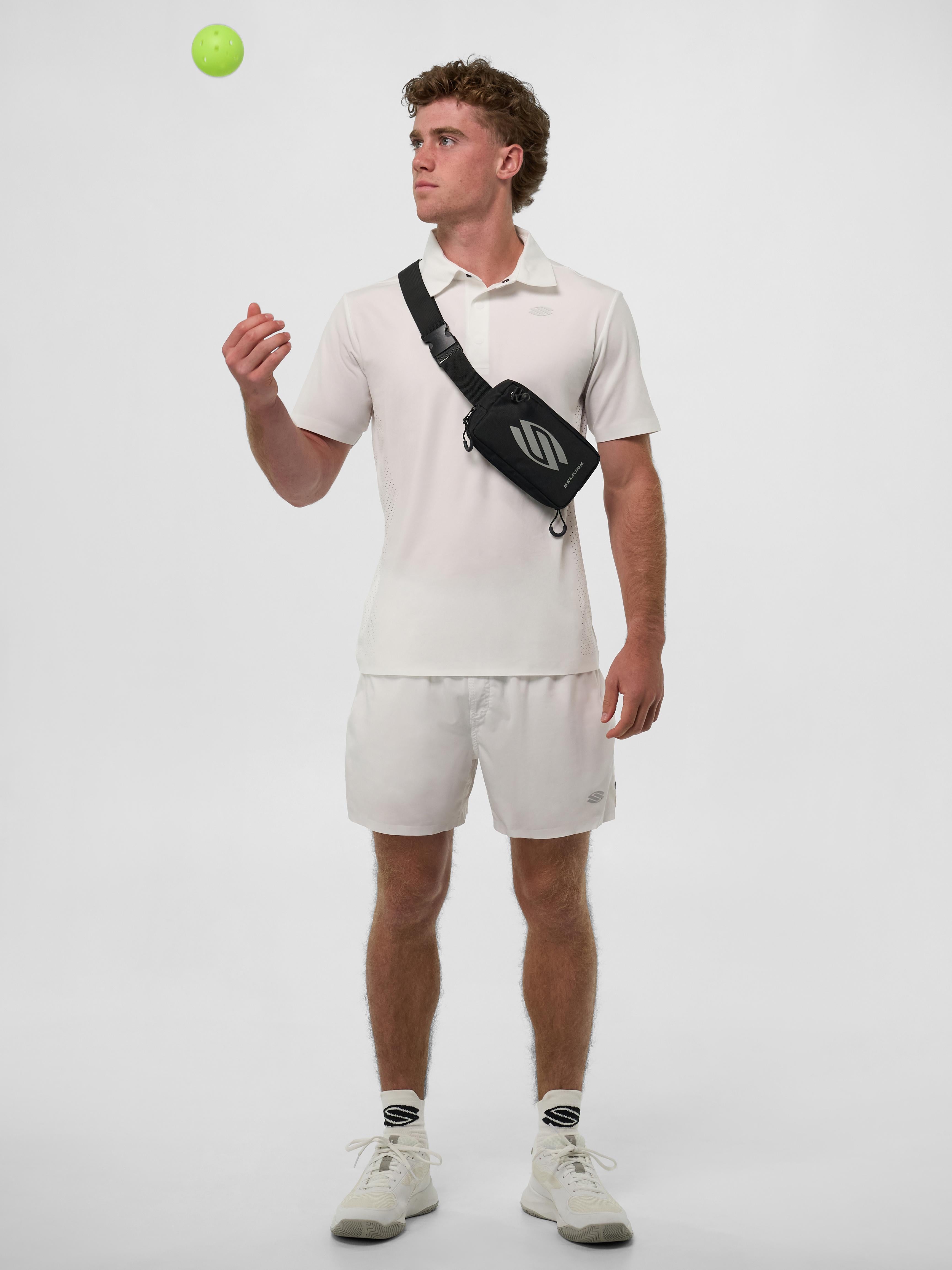 A man wearing a white performance polo and shorts, holding a pickleball, with a black sling bag.