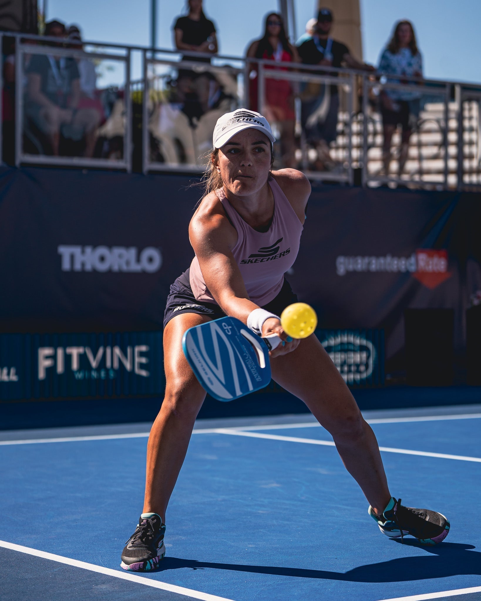 A player in a pink tank top and cap, preparing to hit a pickleball with a paddle on a court.