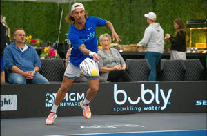 A player in a blue shirt hitting a pickleball with 'bubly sparkling water' logo visible.