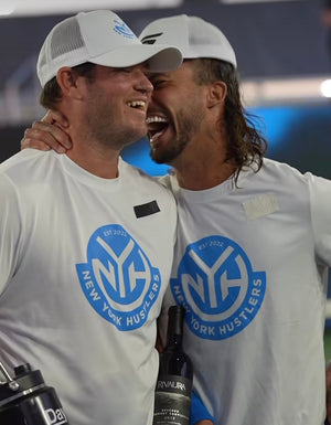 Two players from the New York Hustlers celebrating with a bottle of wine, wearing team shirts.