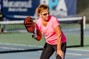 A woman in a pink shirt and sunglasses preparing to hit a pickleball with a paddle on a court.