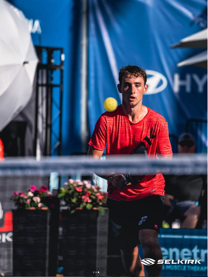 A male player in a red shirt preparing to hit a pickleball during a tournament match.