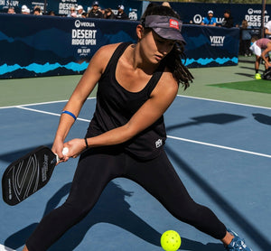A player executing a spin shot in pickleball, wearing a black tank top and cap, with a paddle and ball in play.