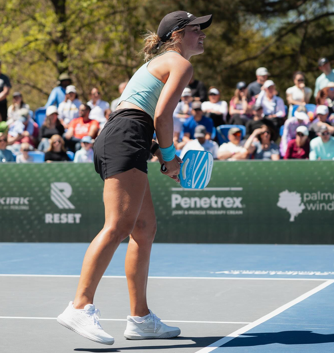 Female pickleball player preparing to serve on outdoor court, wearing light blue tank and black shorts, spectators behind