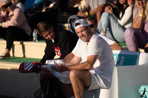 Two pickleball players sit on a sideline bench during a timeout, one wiping his hands and paddle grip with a sweat towel