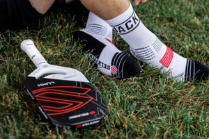 Lower legs wearing white Selkirk crew pickleball socks with “JACK” on the cuff, resting on grass beside a Selkirk LUXX paddle