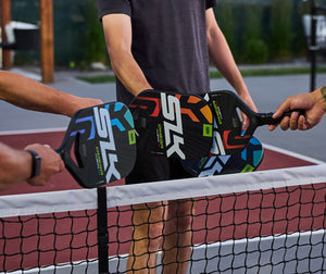 Pickleball players tap their paddles over a net on an outdoor court