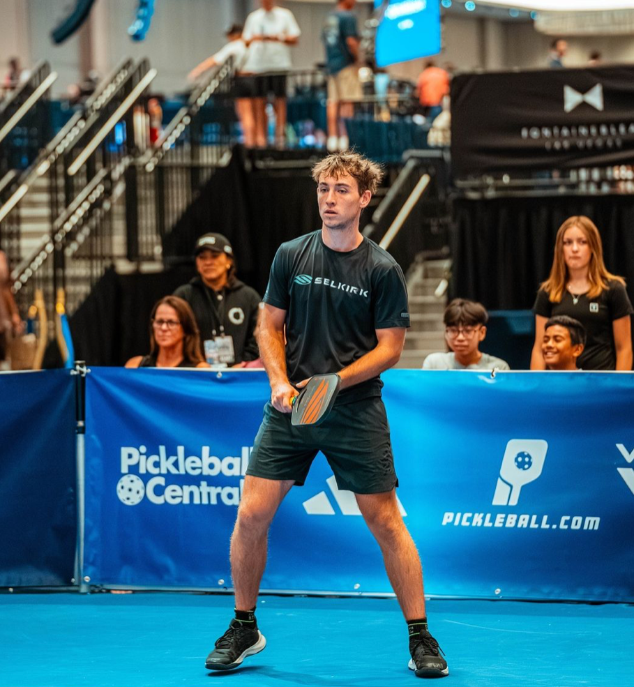 A male pickleball player preparing to serve on a blue court with spectators in the background.