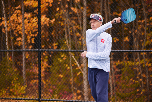 An older man plays pickleball outdoors in long sleeves, with autumn leaves in the background.