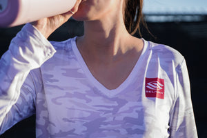 Woman drinking from a water bottle while wearing a lavender camouflage Selkirk shirt