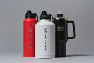 Front view of three Selkirk insulated drinkware: red bottle, white wide-mouth bottle, black tumbler with handle and straw on gray background