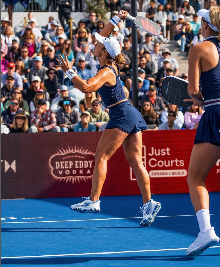 A player in a blue outfit serves during a women's doubles match with spectators in the background.