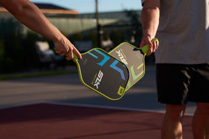 Angled close-up of two Selkirk SLK pickleball paddles showcasing face graphics and neon green trim with an outdoor court background