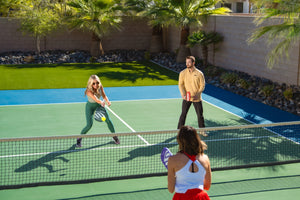 Three pickleball players play on an outdoor court with palm trees and a block wall in the background