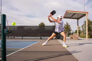 Pickleball player lunging to hit a backhand as a neon yellow ball passes near the net on an outdoor court