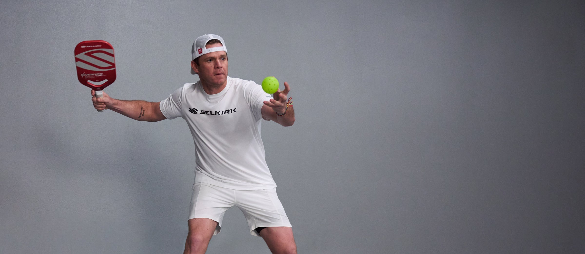 Pickleball player hitting a forehand with a red Selkirk paddle and neon green ball against a gray studio background