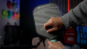 A hand wipes a Selkirk Pickleball paddle with a cloth in a gear-filled room