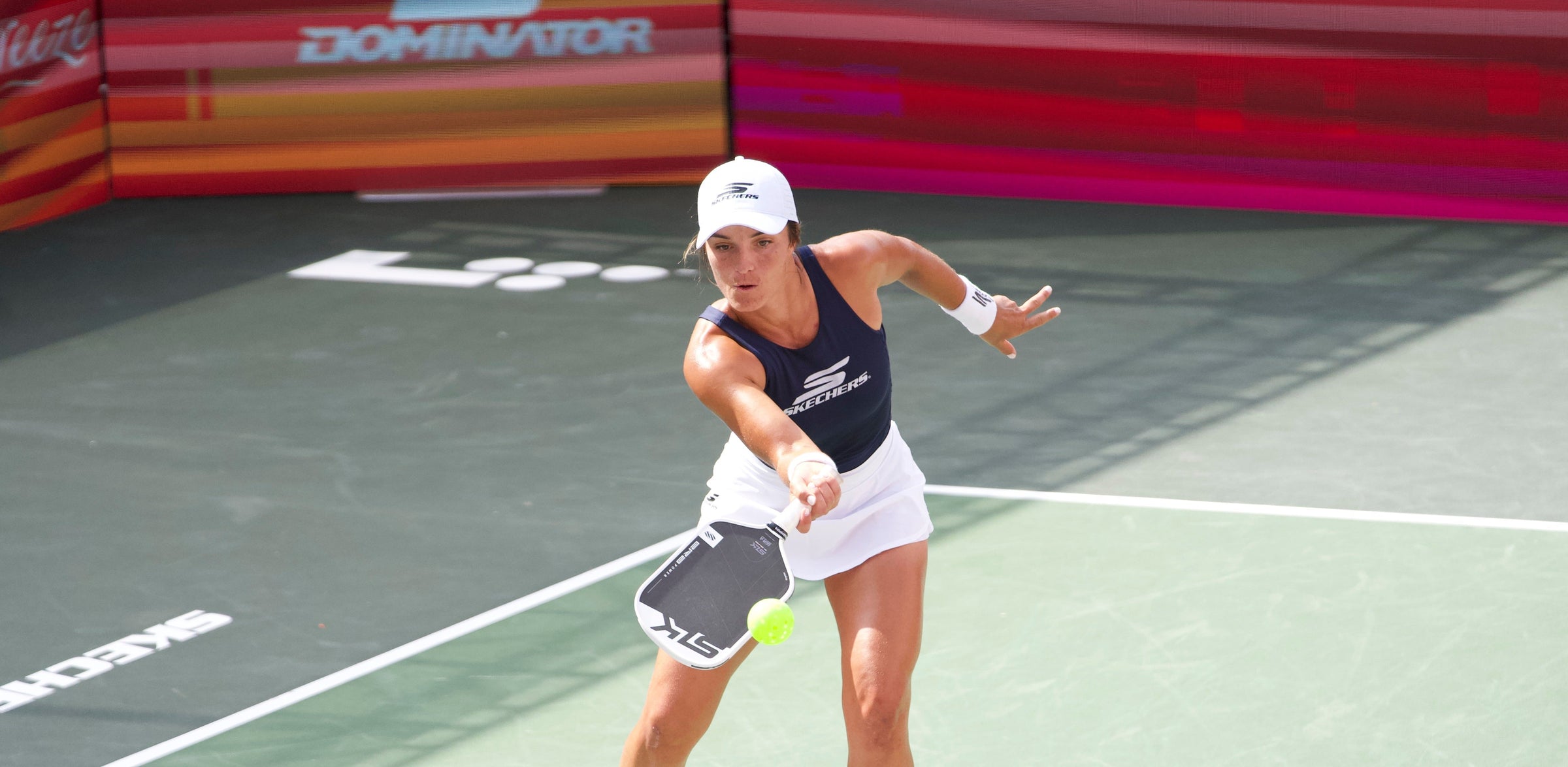 Pickleball player lunging to hit neon ball with paddle, wearing navy tank with SKECHERS logo; DOMINATOR banner behind