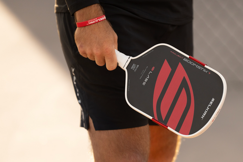 Close-up side view of Project Boomstik pickleball paddle showing black Infinigrit face with red Selkirk logo and white frame while a player holds it on an outdoor court
