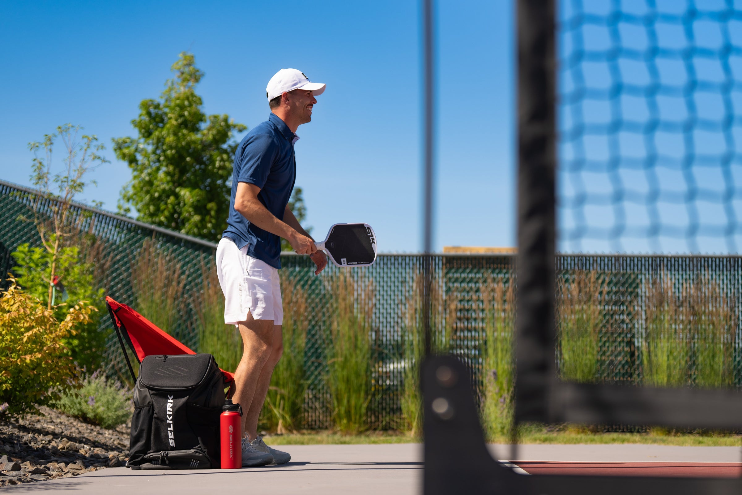 Man in white cap and blue shirt holding a pickleball paddle on an outdoor court; backpack labeled 'SELKIRK' and red bottle at his feet.