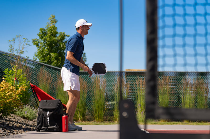 Man in white cap and blue shirt holding a pickleball paddle on an outdoor court; backpack labeled 'SELKIRK' and red bottle at his feet.