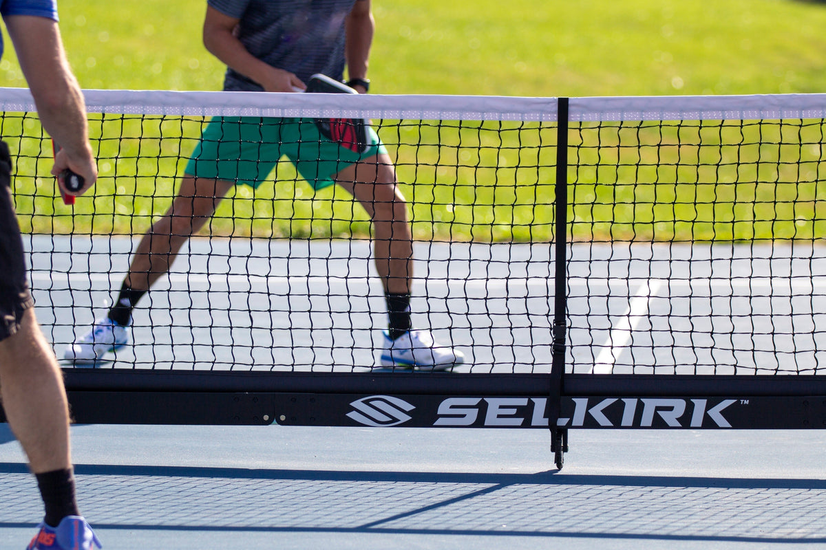 Pickleball players hold paddles on opposite sides of a Selkirk net on an outdoor court