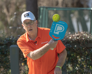 Older man in orange Selkirk polo hitting a yellow pickleball with a blue Selkirk paddle on an outdoor court