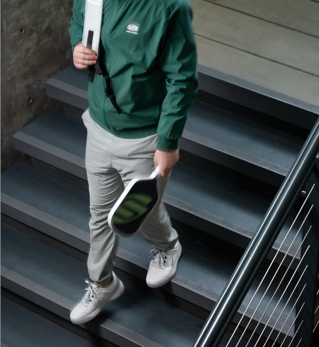Man descends stairs wearing a green Selkirk jacket, white pants, and sneakers while carrying a Pickleball paddle