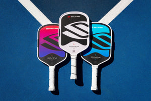 Overhead view of three Selkirk OMNI elongated pickleball paddles in pink, white, and blue on a blue court