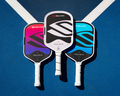 Top down view of three Selkirk OMNI pickleball paddles in pink, white, and blue showcasing large Selkirk logo on a blue court with white lines