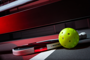 A Boomstik paddle lays on an indoor court next to a pickleball. 
