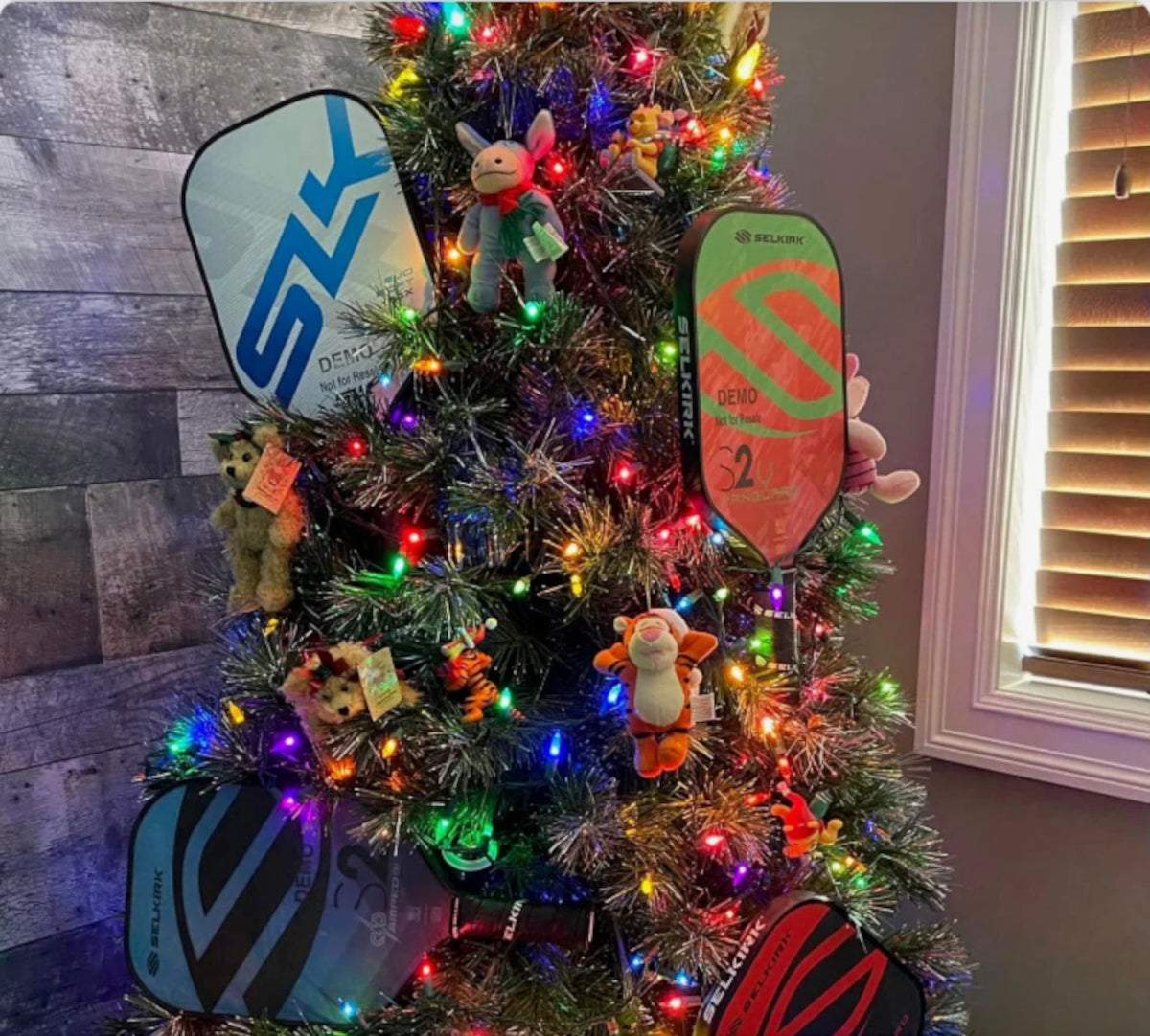 A Christmas tree decorated with colorful lights and four Selkirk pickleball paddles among ornaments.