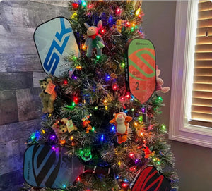A Christmas tree decorated with colorful lights and four Selkirk pickleball paddles among ornaments.