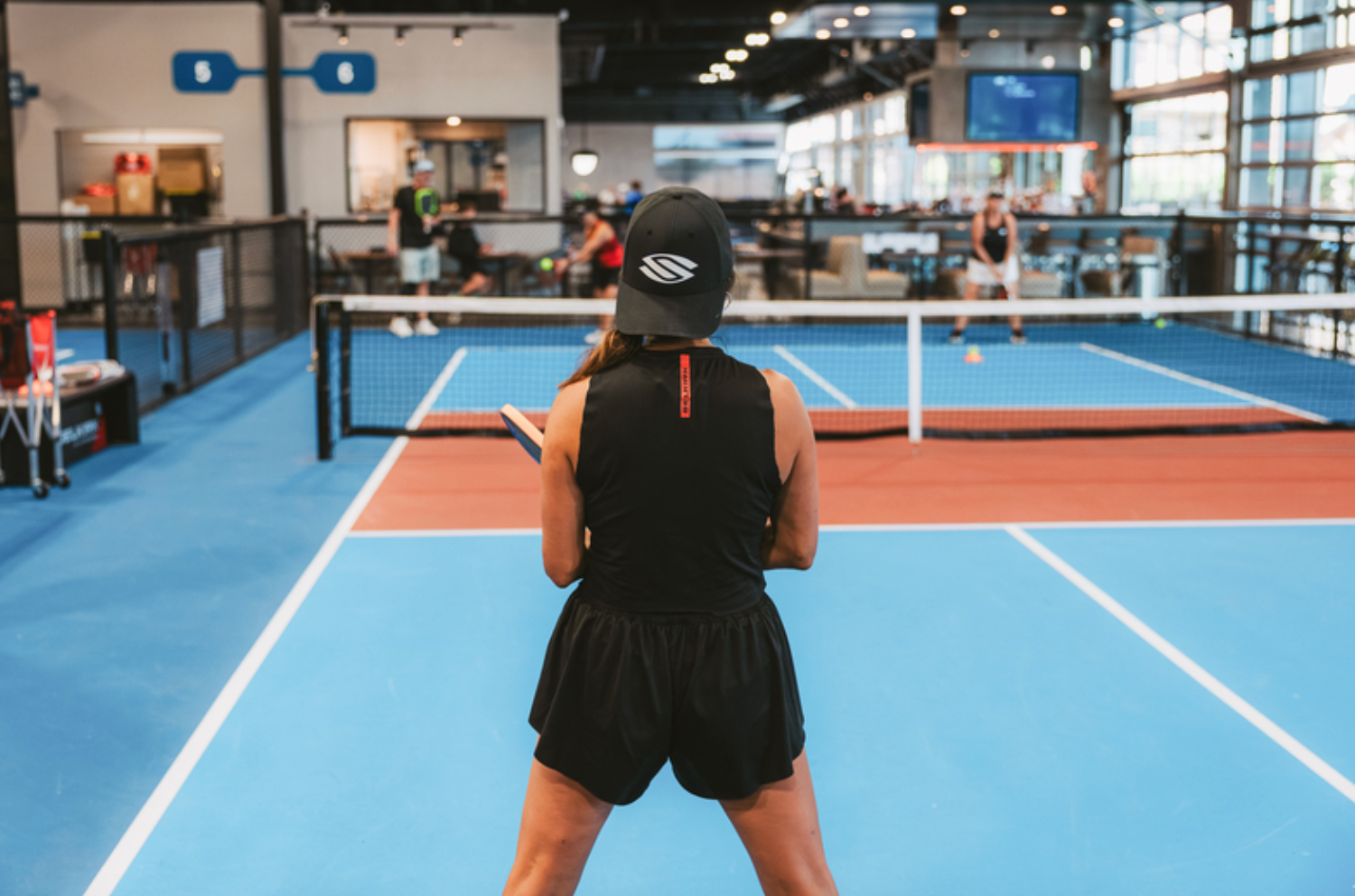 An image shows a woman's back as she prepares to receive a pickleball serve on an indoor court. 