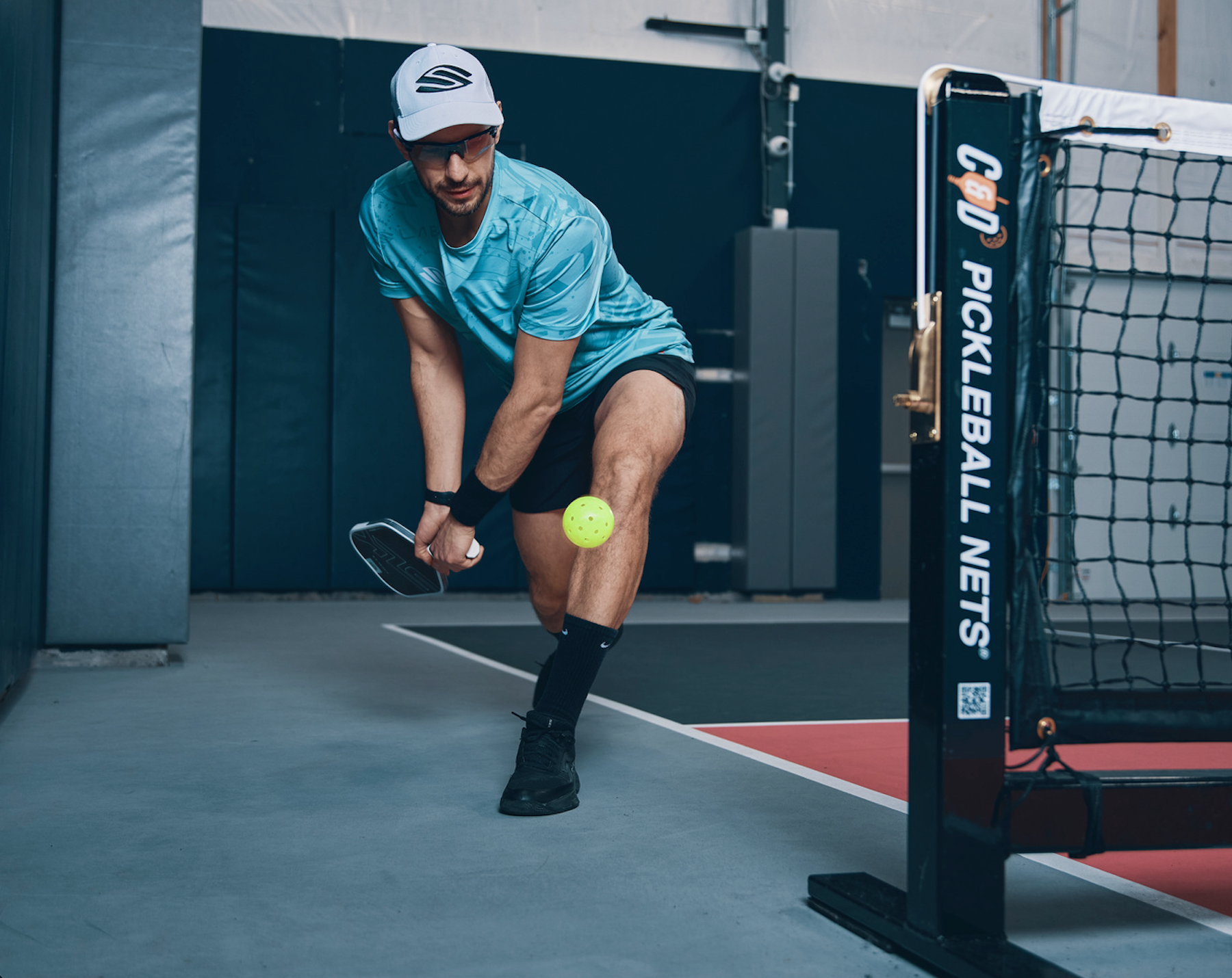 A man attempts to hit an around-the-post shot on an indoor pickleball court. 