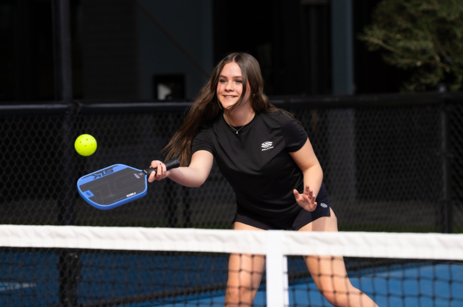 A teenage girl smiles as she hits a volley dink with her SLK Neo pickleball paddle. 