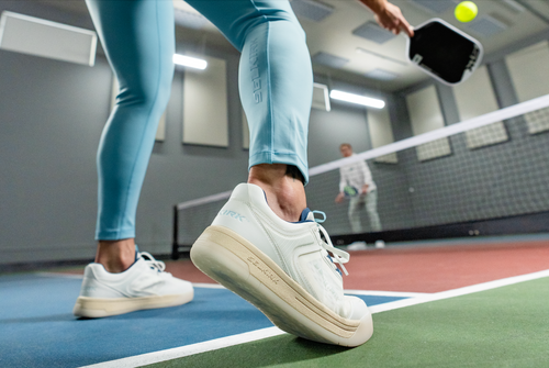 A close-up shows a woman's foot wearing the Legacy Pro pickleball shoes while she plays.