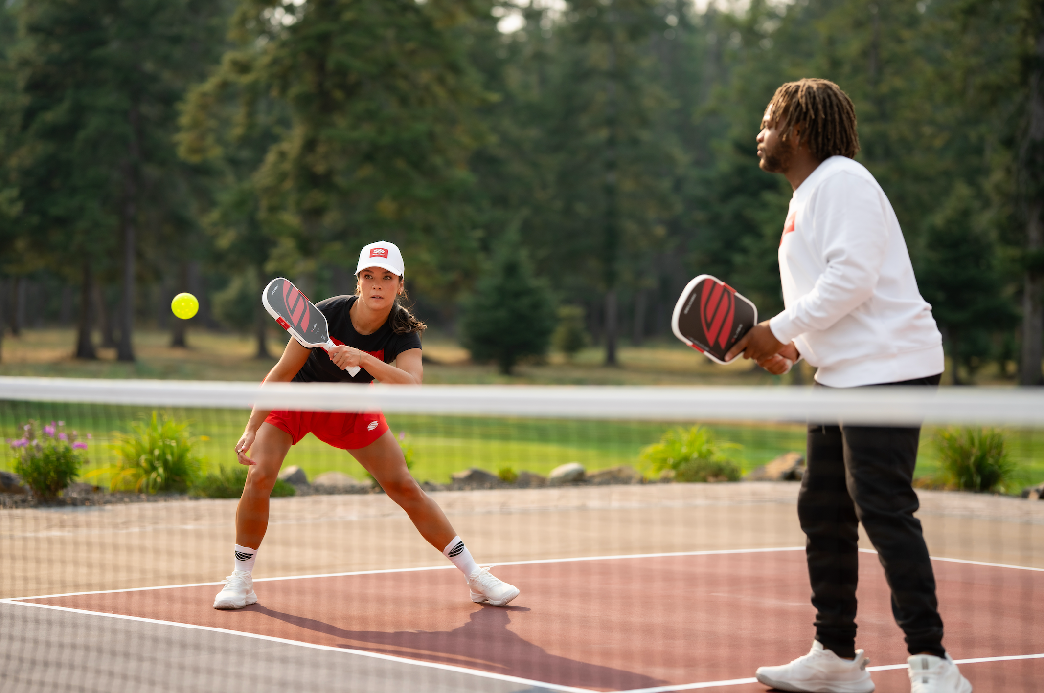 A man talks to a woman as she hits a volley on an outdoor pickleball court. 