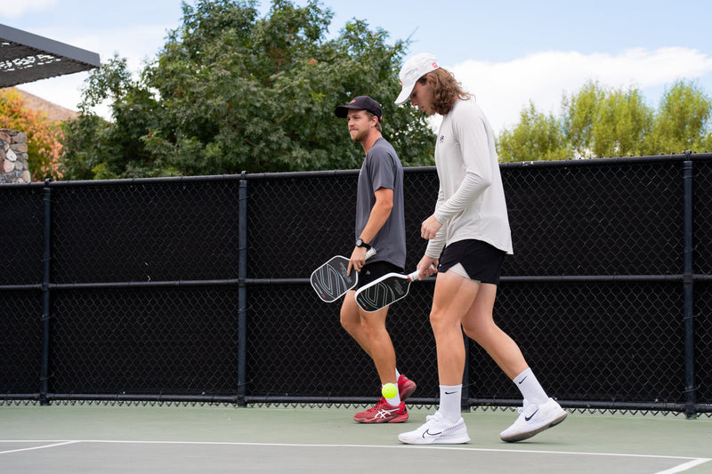 Two pickleball players wear white, lightweight breathable clothing to play outside. 
