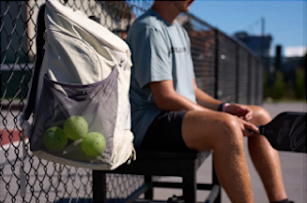 A white Selkirk pickleball backpack clipped to a fence with green pickleballs visible in the mesh pocket.
