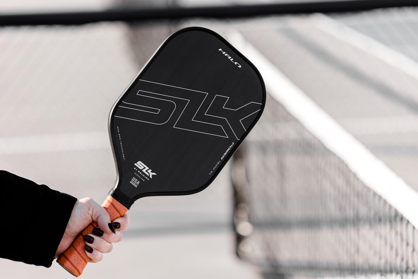 A person holding a Selkirk SLK HALO pickleball paddle with a black surface and orange grip.