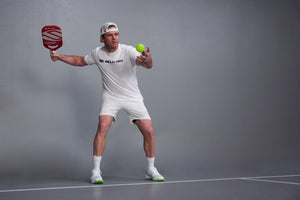 Pickleball player in white SELKIRK shirt serving with red paddle and bright green ball on indoor court.