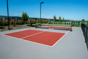 Portable black net across an outdoor court with maroon surface, yellow lines, chain-link fence and trees.