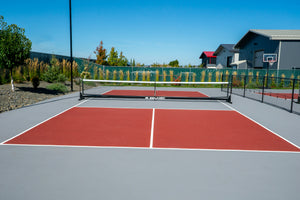 Outdoor red-and-gray pickleball court with a SELKIRK net, chain-link fence, houses and a basketball hoop in background.