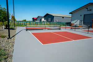 Outdoor pickleball courts with red surfaces, two nets, chain-link fences, and nearby storage buildings under clear blue sky.